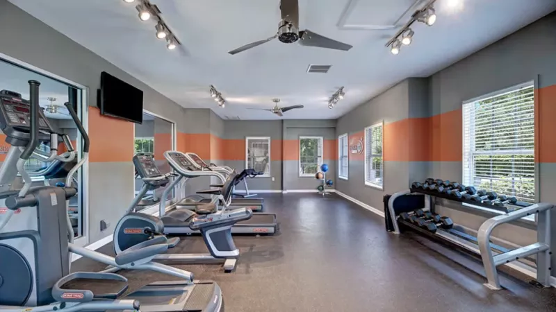 A gym interior featuring various exercise equipment including treadmills, exercise machines, and open space on the floor. The room has a ceiling and walls enclosing the area.