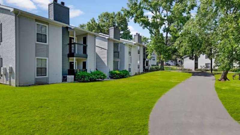 Apartment building surrounded by trees and grass, set against a clear sky with a few clouds. The property features a well-manicured lawn and landscaping, with visible windows and a driveway.