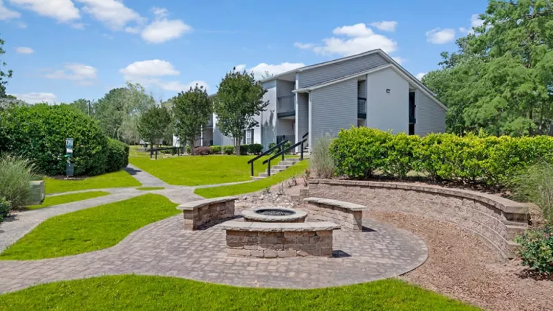 A building with a fountain in front, surrounded by grass and landscaping, with trees and a clear sky above.