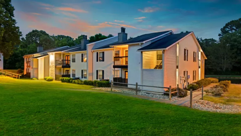 A building with a fountain in front, surrounded by grass and landscaping, with trees and a clear sky above.