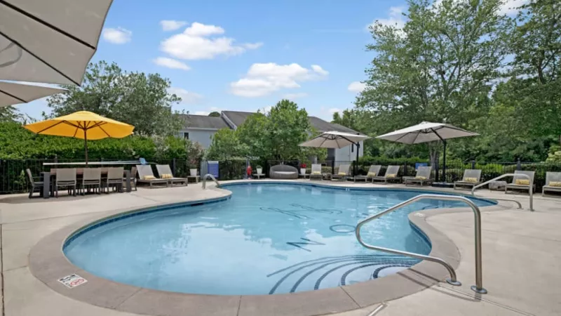 A swimming pool surrounded by umbrellas, with trees and a clear sky in the background, creating a relaxing outdoor atmosphere at an apartment building.