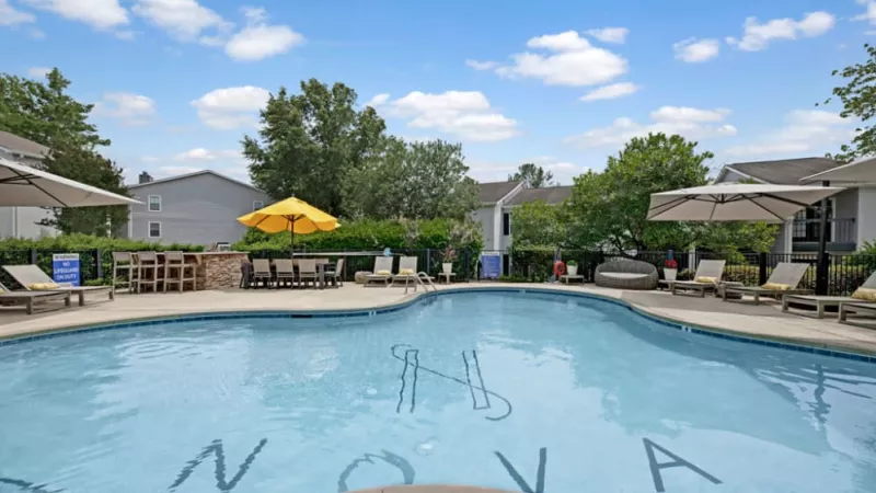 A swimming pool with a sign that reads "NOVA" surrounded by trees, under a blue sky with clouds, located in a resort area.