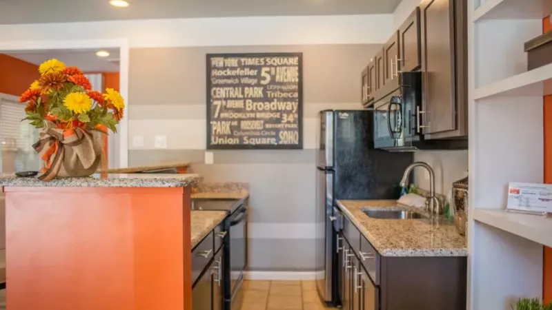 A spacious room featuring several tables and chairs arranged throughout the space, with designs that suggest a kitchen or dining area. The decor includes a studio couch and coffee table, complemented by warm orange tones.