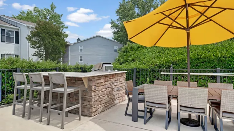 A patio featuring several chairs arranged around a table, with an umbrella providing shade. Lush greenery and a plant are visible nearby, creating a relaxing outdoor space ideal for summer gatherings.