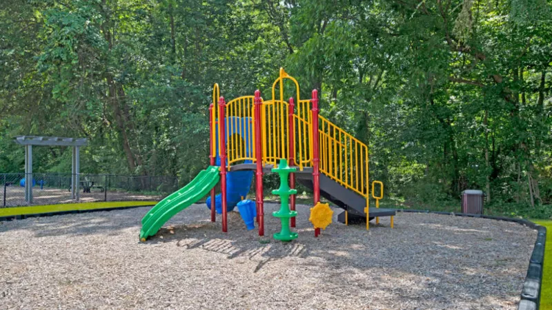 A playground featuring various play equipment such as slides and a jungle gym, surrounded by trees in the background. The ground is set up for outdoor play in a public park space.