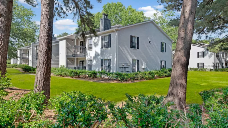 White Apartment homes with a green lawn surrounded by trees and a clear sky.