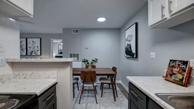A kitchen featuring a table with chairs, surrounded by cabinetry and countertops. The space includes a sink, oven, microwave, and other kitchen appliances, with a floor and ceiling visible in the room's interior design.