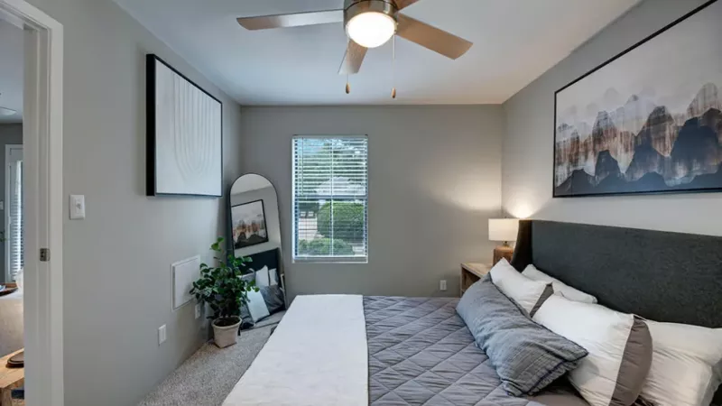 A cozy bedroom featuring a bed with pillows and linens, a ceiling fan overhead, wall decor, a nightstand with a lamp, and window treatments allowing natural light to enter. The room has a warm interior design aesthetic.
