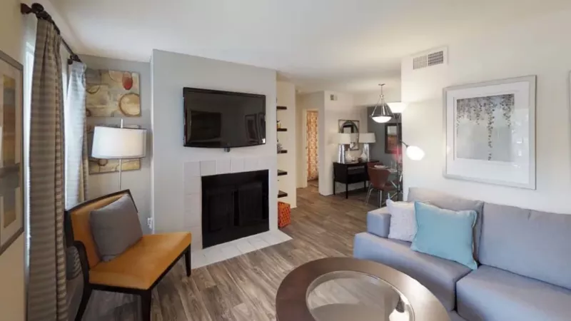 A cozy living room featuring a fireplace, adorned with a sofa and loveseat. The space includes a coffee table, flat-screen television, and various decorative pillows. The flooring is a mix of wood and laminate, complemented by cabinetry on the walls. Large windows allow natural light to fill the room.