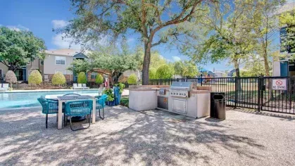 Outdoor grilling station and dining area set against a serene backdrop of lush greenery and a sparkling pool. Featuring modern grills, a shaded dining table, and a relaxing atmosphere, this space is perfect for outdoor entertaining.