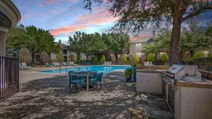 Evening view of a serene poolside area featuring a modern grilling station, outdoor seating, and lush landscaping. The warm glow of sunset creates a tranquil and inviting ambiance for outdoor gatherings.