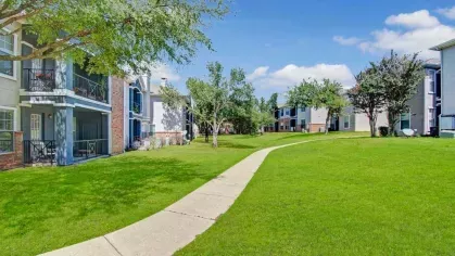 Beautifully landscaped community grounds with spacious green lawns, walking paths, and mature trees. The tranquil setting is complemented by nearby modern apartment buildings with private balconies.