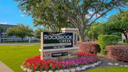 A welcoming exterior sign for "Rockbrook Creek Apartment Homes" with landscaped flowers.