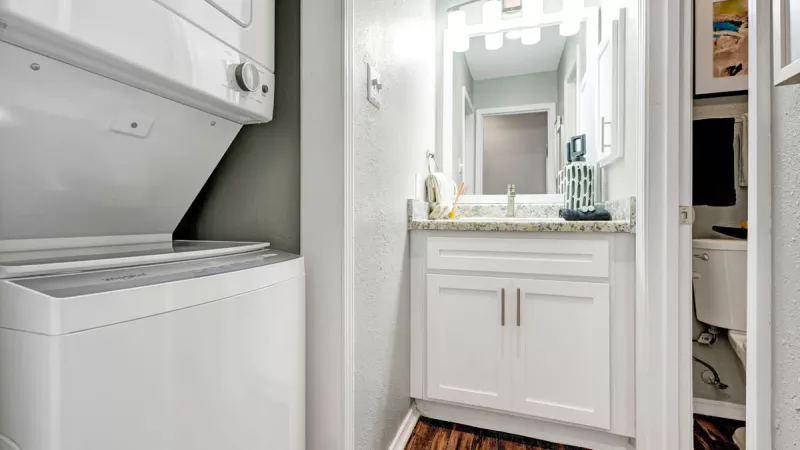 Compact and functional laundry area adjacent to a bright bathroom with a sleek white vanity, modern fixtures, and a large mirror. The space features wood-style flooring and thoughtful design for convenience and efficiency.
