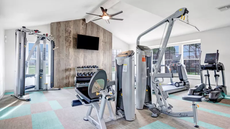 Modern fitness center featuring a variety of exercise equipment, including strength training machines, cardio machines, and free weights. The space is bright and airy, with large windows, light flooring, and a mounted TV for an enjoyable workout environment.