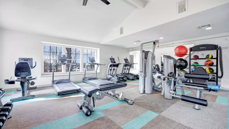 Bright and spacious fitness center equipped with modern exercise machines, including treadmills, ellipticals, and strength-training equipment. The space features large windows, colorful flooring, and ceiling fans, creating an inviting and energizing workout environment.
