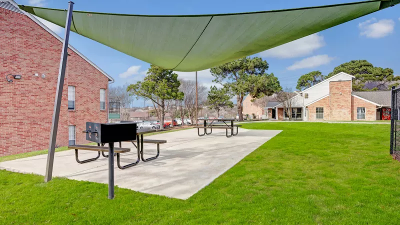  Outdoor picnic area featuring shaded seating with picnic tables, a barbecue grill, and lush green grass. The space is ideal for gatherings and relaxation, surrounded by a bright blue sky and community buildings.
