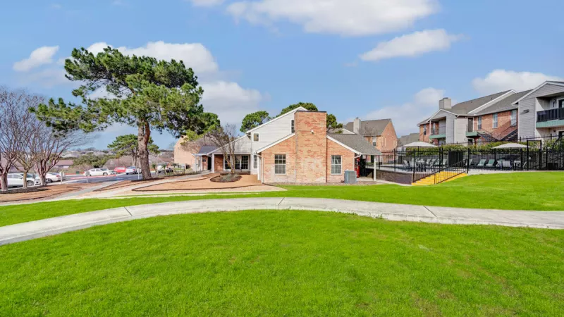 Expansive green lawn with a well-maintained pathway, leading to modern apartment buildings in the background. The open space is perfect for outdoor activities, surrounded by trees and a bright blue sky.