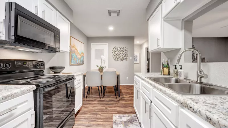  Modern galley-style kitchen featuring sleek white cabinetry, stainless steel appliances, and wood-style flooring. The space opens to a dining area, with bright lighting and clean finishes creating a stylish and functional atmosphere.