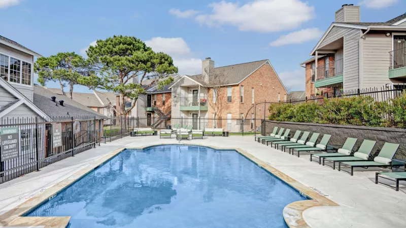  Refreshing outdoor pool with lounge chairs surrounding the clear blue water, set against a backdrop of modern apartment buildings and green trees. The bright blue sky enhances the relaxing atmosphere of the pool area.
