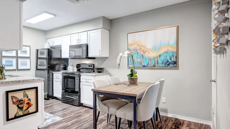  Modern kitchen and dining area featuring white cabinetry, stainless steel appliances, and wood-style flooring. A cozy dining table with upholstered chairs sits against a stylish accent wall with colorful artwork, creating a bright and inviting atmosphere.