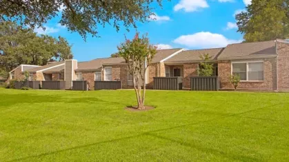 A row of single-story brick apartment homes surrounded by a well-maintained green lawn and shaded by mature trees.