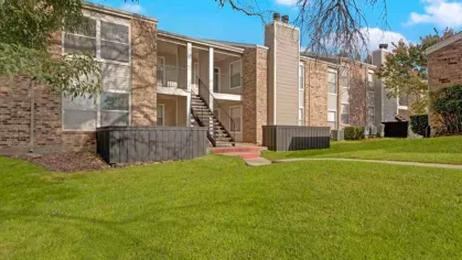 A view of a brick apartment complex with a central staircase and open balconies, surrounded by lush greenery.