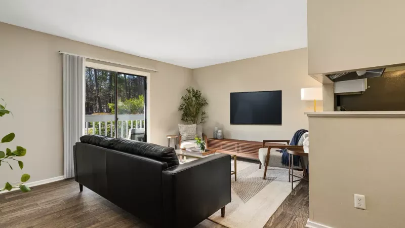 A modern living room with a black leather sofa, a flat-screen TV mounted on the wall, and a large sliding glass door leading to an outdoor space.