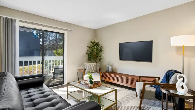 A bright and cozy living room featuring a black leather sofa, glass coffee table, and a large window with vertical blinds.