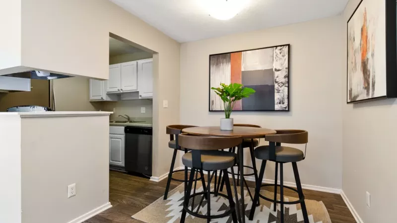A compact dining area with a round wooden table and four chairs, next to an open kitchen with white cabinetry.
