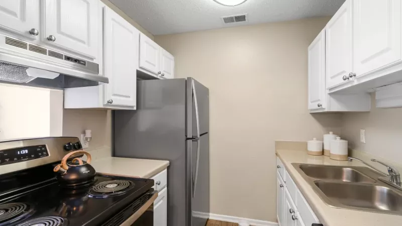 A narrow galley kitchen with white cabinets, a stainless steel refrigerator, and a black electric stove.