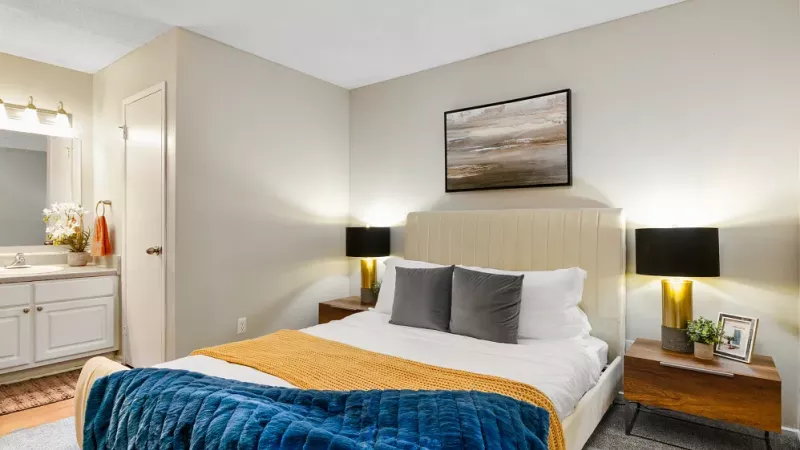 A cozy bedroom with a beige upholstered bed, colorful throw blankets, and matching nightstands with table lamps.