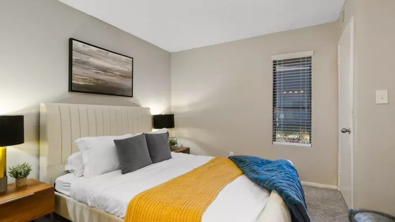 A bright bedroom with a neatly made bed, accented with a mustard-yellow blanket and modern bedside lighting.