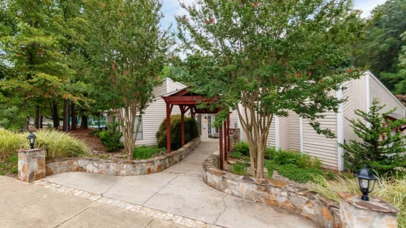 A stone pathway leading to a charming clubhouse surrounded by landscaped greenery and mature trees.
