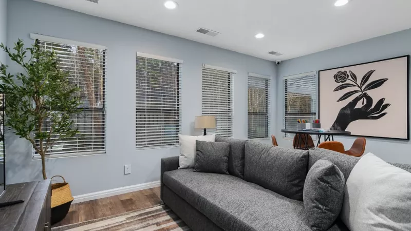 A bright, airy lounge featuring a gray sectional sofa, artistic wall decor, and a workspace area with natural light from multiple windows.