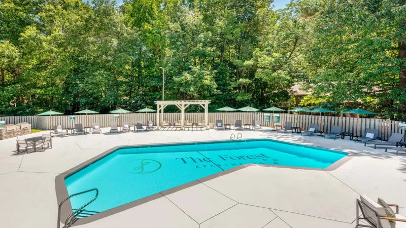 A bright outdoor pool area surrounded by lounge chairs, green umbrellas, and a shaded pergola, located at The Forest Apartments, with a backdrop of lush greenery.