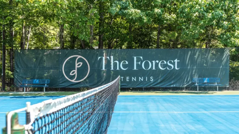 A tennis court with "The Forest Tennis" banner displayed on a green backdrop, surrounded by trees.