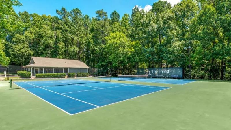 A vibrant blue tennis court with wooded surroundings and a nearby clubhouse.