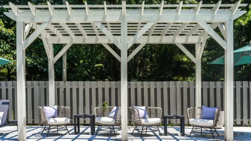 An outdoor seating area under a white pergola, featuring comfortable chairs with cushions and side tables, surrounded by a wooden fence and greenery.