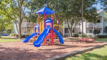 A vibrant children's playground with colorful slides and climbing structures surrounded by green trees and apartment buildings in the background.