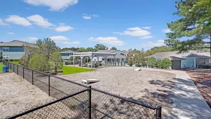 A spacious fenced-in outdoor area with a clear pathway leading to picnic tables and a pergola, surrounded by greenery and a residential complex.