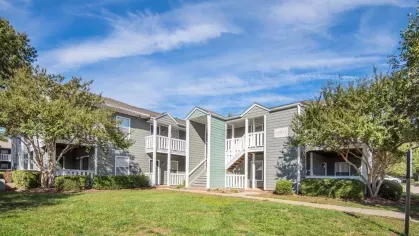 Two-story apartment buildings with balconies surrounded by lush greenery and a bright blue sky.