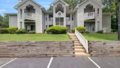 A central entrance leading to gray apartments with balcony railings and manicured bushes.