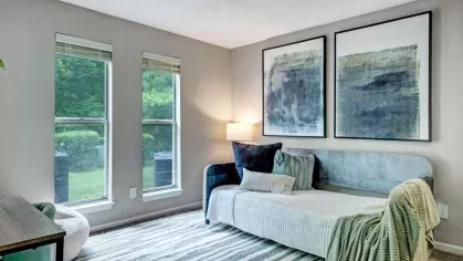 A bright and airy room featuring a cozy daybed, decorative artwork, and large windows overlooking greenery.