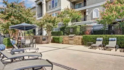 Outdoor lounging area with reclining chairs, a dining table, and lush landscaping in a modern apartment complex.