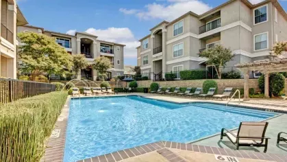 Sparkling swimming pool surrounded by apartment buildings, trees, and lounge chairs under the bright blue sky.