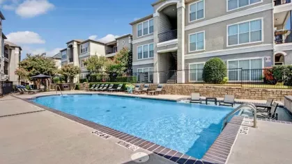 A rectangular swimming pool surrounded by lounge chairs, green hedges, and contemporary apartment buildings.