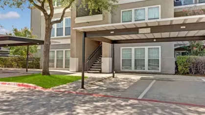 Covered parking spaces alongside a residential building with landscaped greenery and a shaded tree.