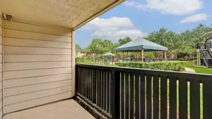 A private balcony with wood siding and a railing, offering a view of a landscaped community area with gazebos and greenery.