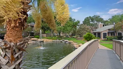 A scenic pond with a fountain, surrounded by lush greenery and accessed by a charming wooden bridge.